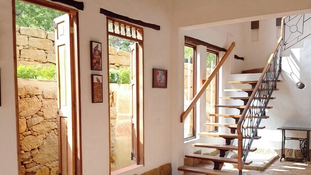 A spacious interior with a stone wall, a wooden staircase, and a view of greenery outside. - Color Marrón (Barichara)
