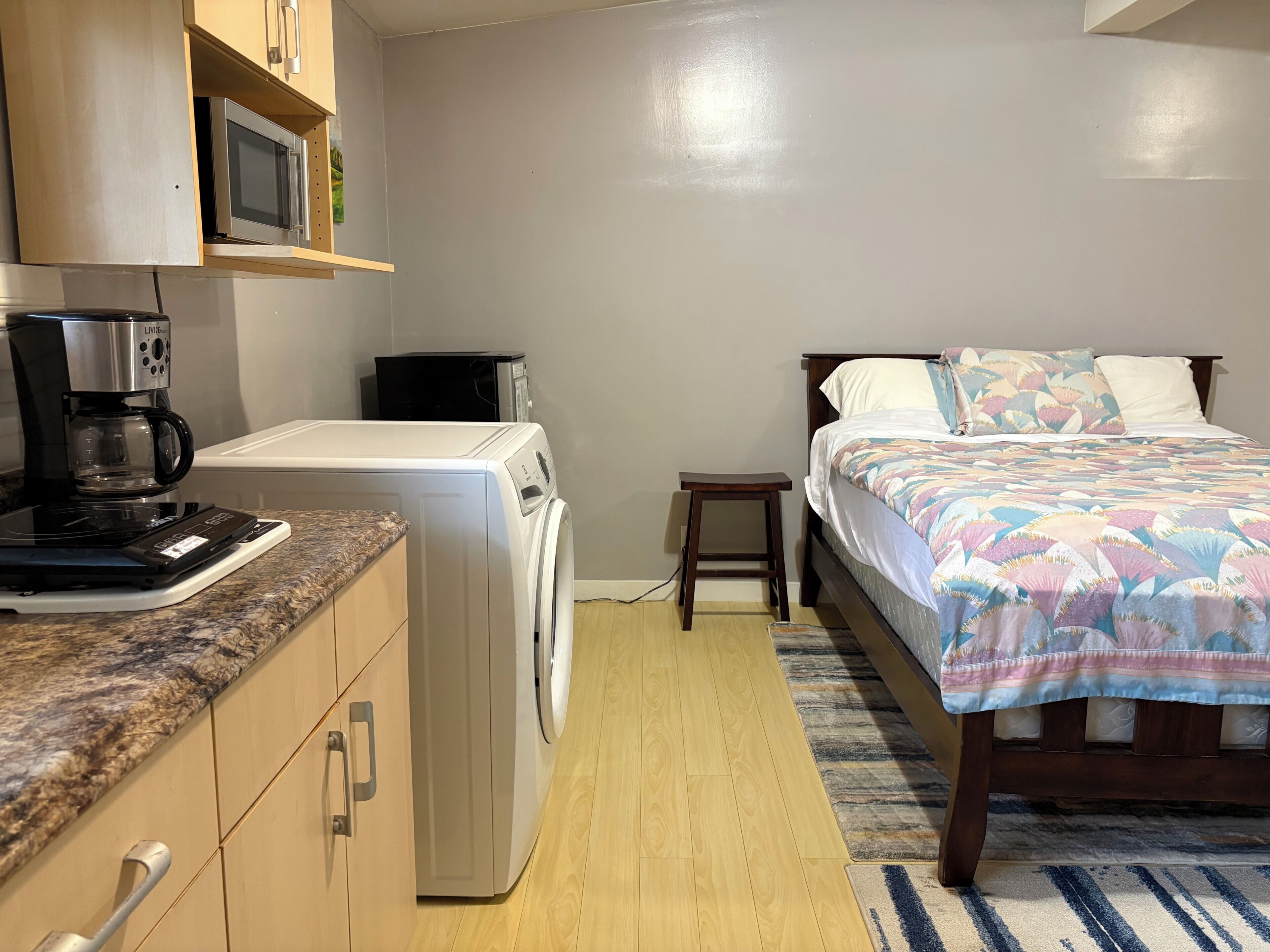 A compact bedroom with a bed, a washing machine, a coffee maker, and a microwave.
