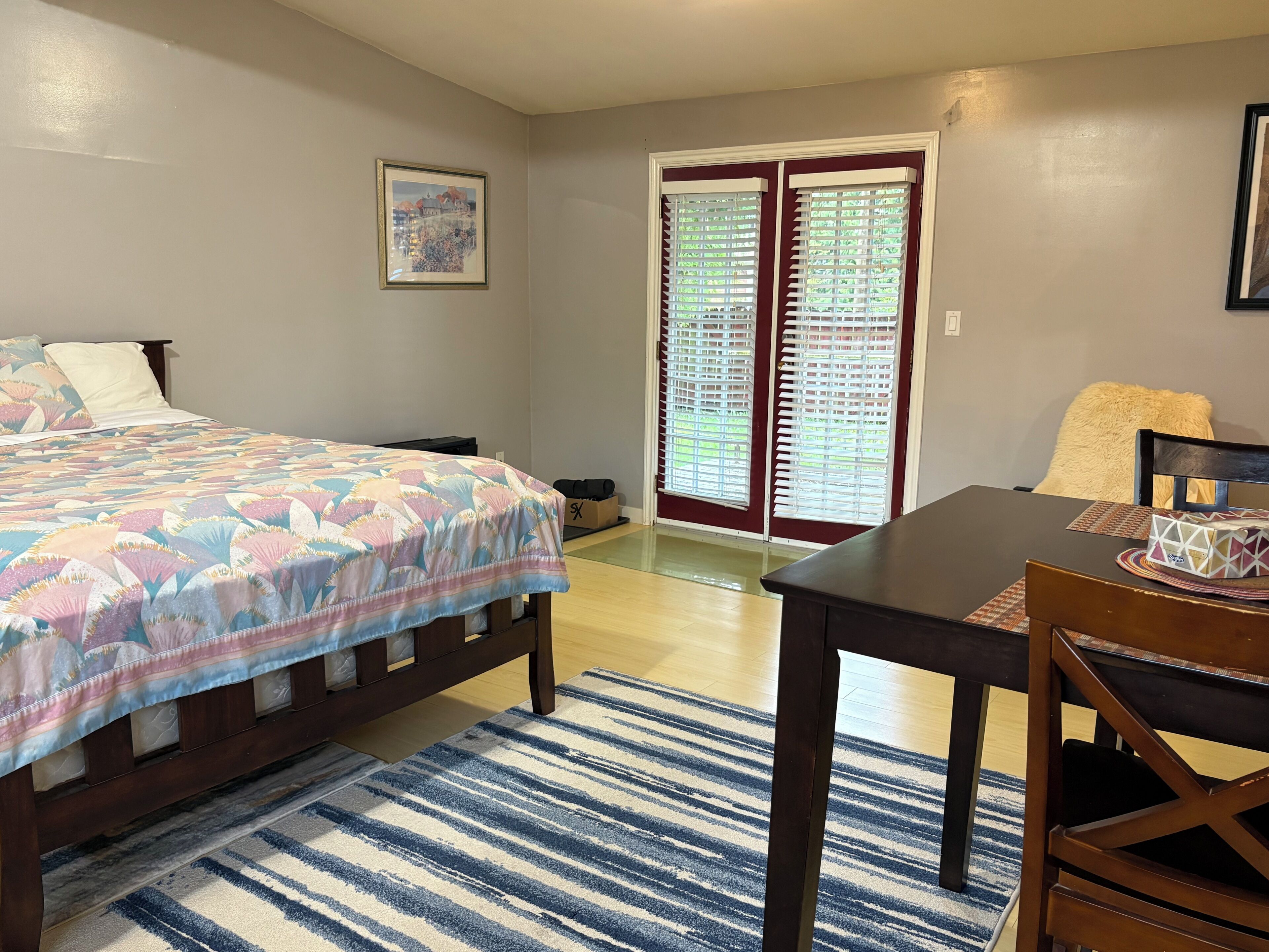 A bedroom with a bed, a desk, a chair, a window with blinds, a door, and a framed picture on the wall.