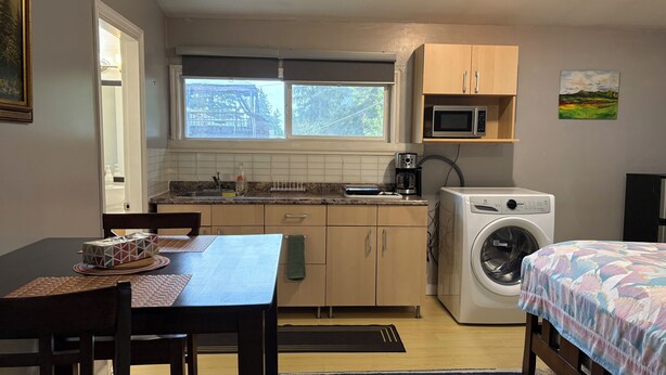 A compact kitchen with a microwave, coffee maker, and washing machine. There is a dining table with chairs and a bed with a colorful bedspread.