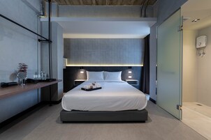 Una habitación de hotel moderna con una cama grande, mesitas de noche, un pequeño escritorio y un muro divisor de vidrio.