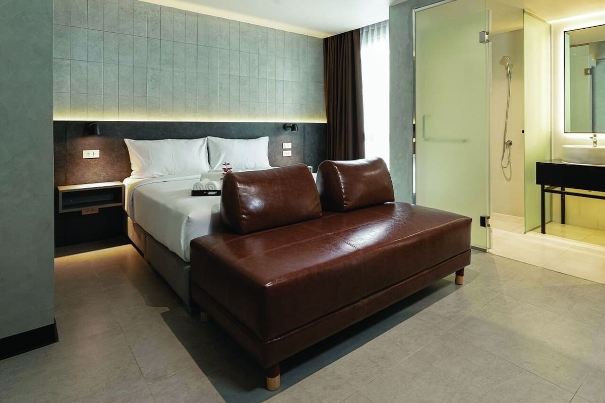 Quarto de hotel moderno com uma cama, um sofá de couro marrom, uma mesinha de cabeceira, um espelho e uma área de chuveiro.