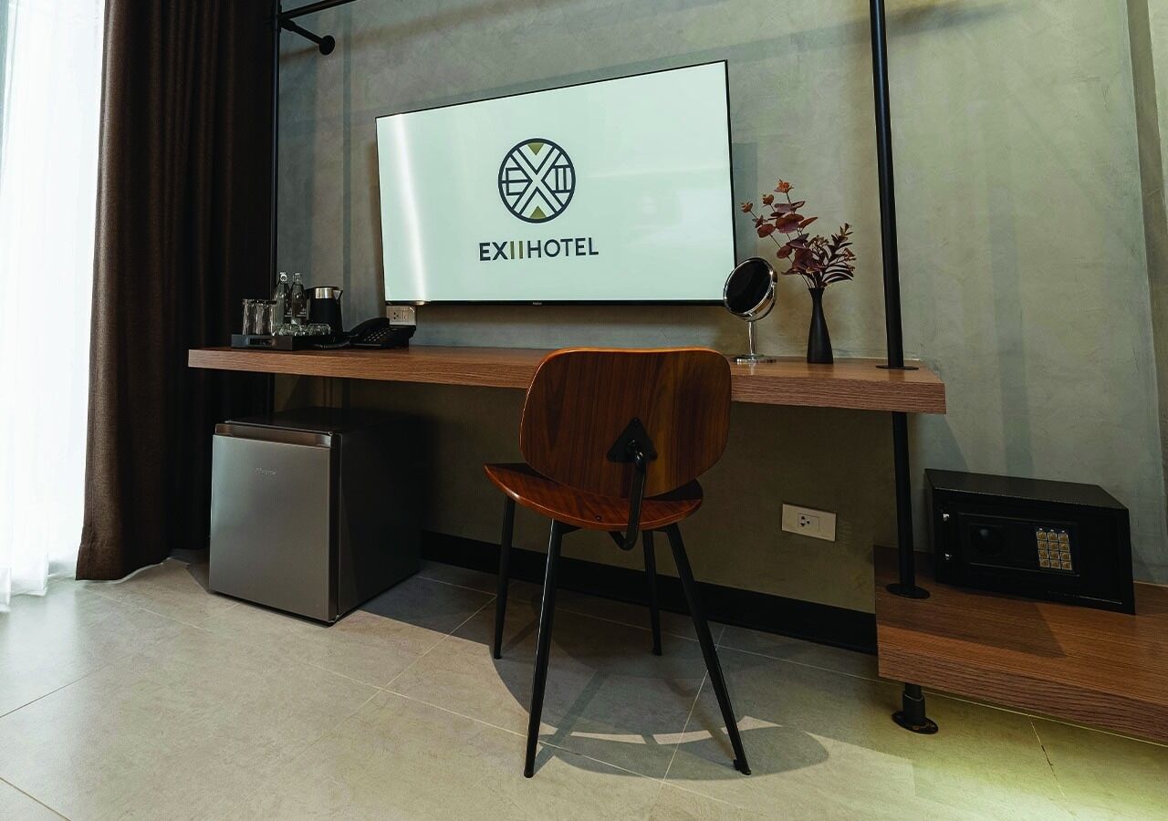 A modern hotel room with a flat-screen TV mounted on the wall, a wooden desk with a chair, a small refrigerator, and a safe on the floor.