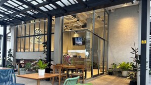 An outdoor seating area with tables and chairs, potted plants, and a glass wall with a sign reading "HOTEL". - EXII Hotel Ngamwongwan (Nonthaburi)