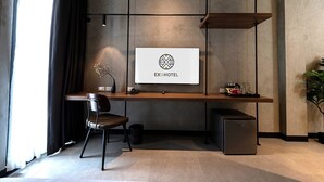 A modern hotel room with a minimalist design, featuring a desk with a computer, a chair, a small refrigerator, and a large window with curtains. - EXII Hotel Ngamwongwan (Nonthaburi)