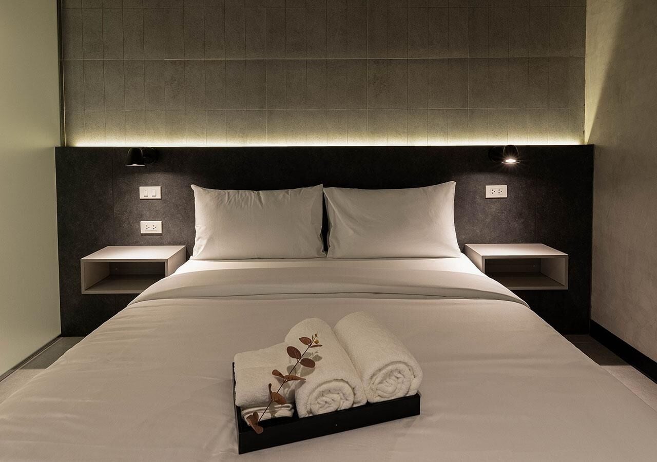 Un lit bien fait, avec du linge de lit blanc, et un plateau contenant des serviettes pliées. Il y a deux tables de chevet, chacune avec une lampe, de chaque côté du lit.