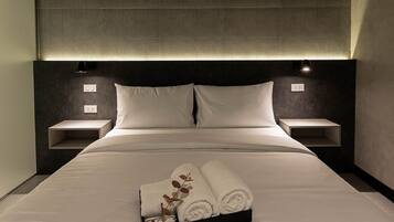Un lit bien fait, avec du linge de lit blanc, et un plateau contenant des serviettes pliées. Il y a deux tables de chevet, chacune avec une lampe, de chaque côté du lit.