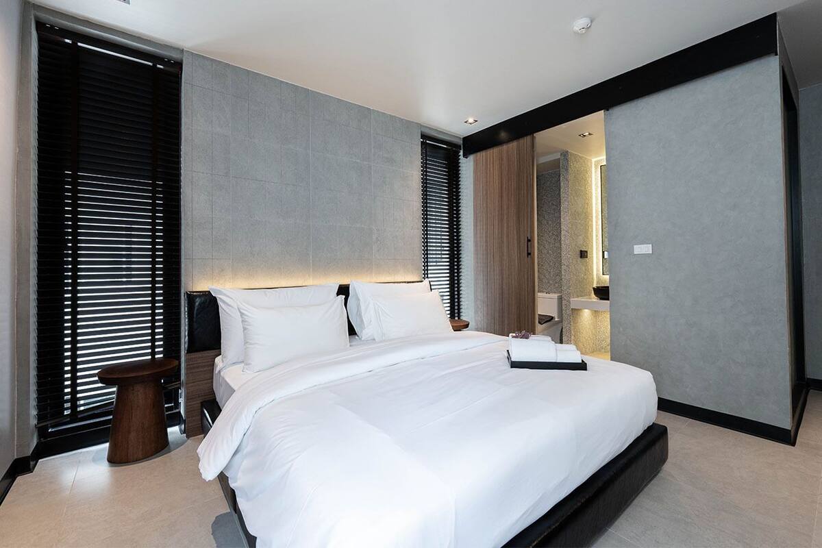 Quarto de hotel moderno com uma cama grande, uma mesinha de apoio e um banheiro visível através de uma porta de correr.