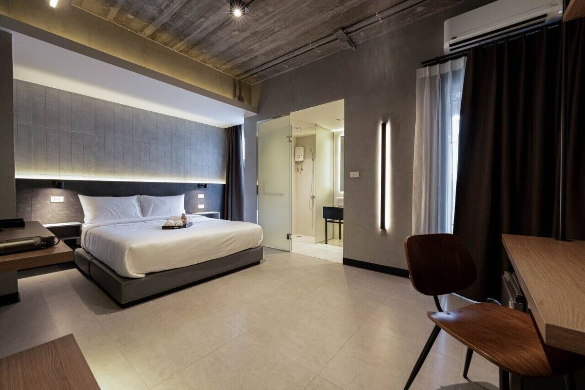 Quarto de hotel moderno com uma cama grande, uma escrivaninha e uma cadeira. Há uma porta de vidro que dá para o banheiro.