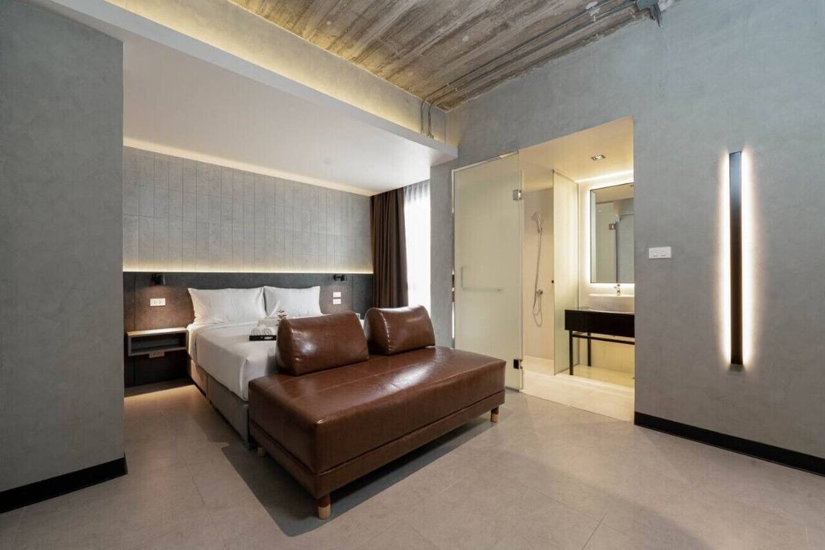 Quarto de hotel moderno com uma cama grande, uma mesa de cabeceira, um sofá e um banheiro visível por uma porta aberta.