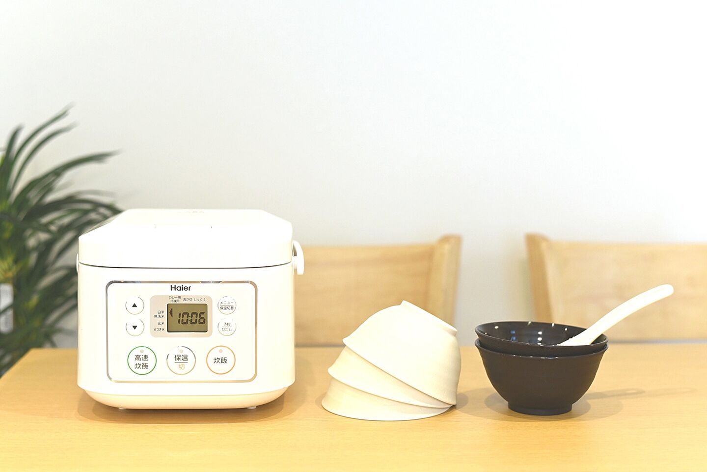 A white rice cooker with a digital display, a white cloth, and a black bowl with a white spoon on a wooden table.