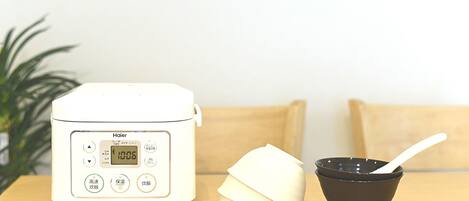 A white rice cooker with a digital display, a white cloth, and a black bowl with a white spoon on a wooden table.