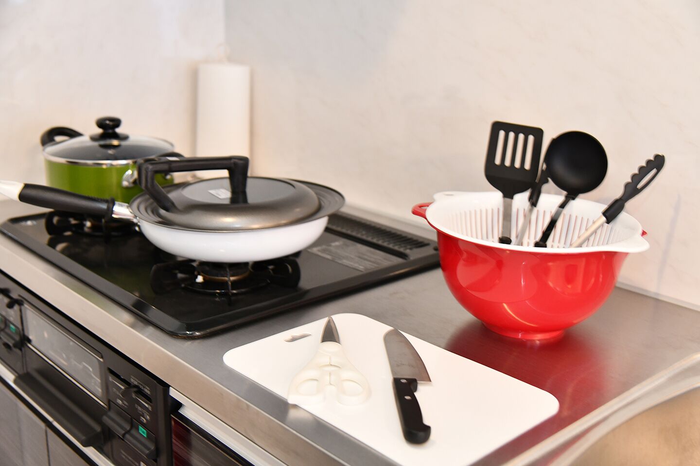 A kitchen with a stove, a frying pan, a red colander with utensils, and a white cutting board with a knife.