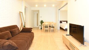 A modern living room with a brown sofa, a wooden dining table with chairs, a flat-screen TV, and a bed in the background. - GLASS YAMAHANA EAST (Sapporo)