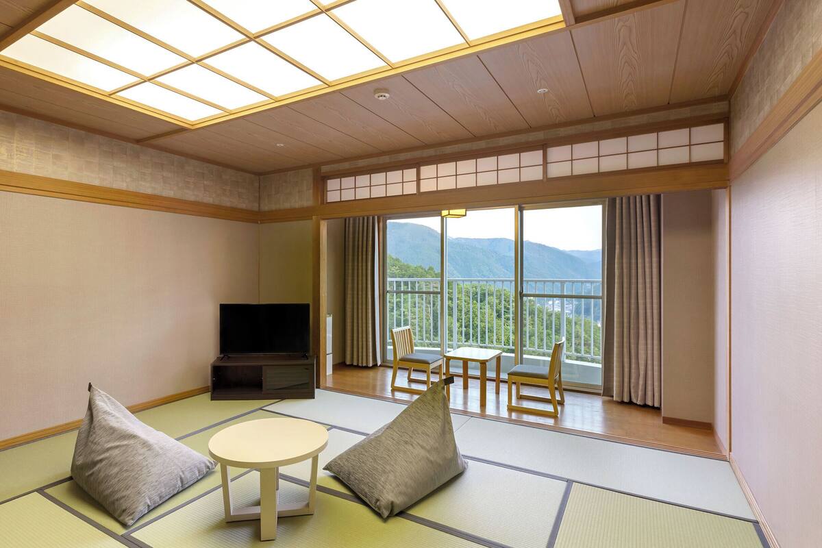 A traditional Japanese-style room with tatami flooring, a television, a table with chairs, and a balcony with a mountain view.