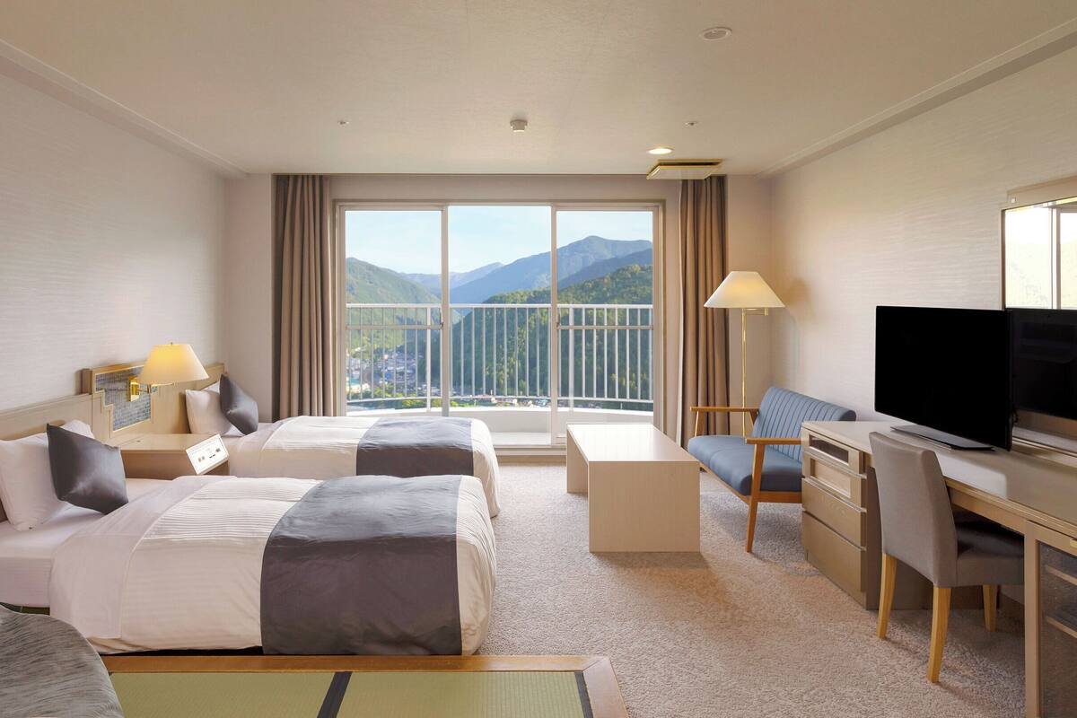 A hotel room with two beds, a desk, a chair, a TV, and a balcony with a mountain view.
