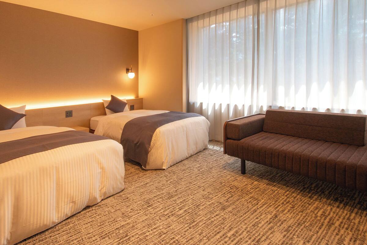 A hotel room with two beds, a sofa, a large window with sheer curtains, and a wall-mounted light fixture.