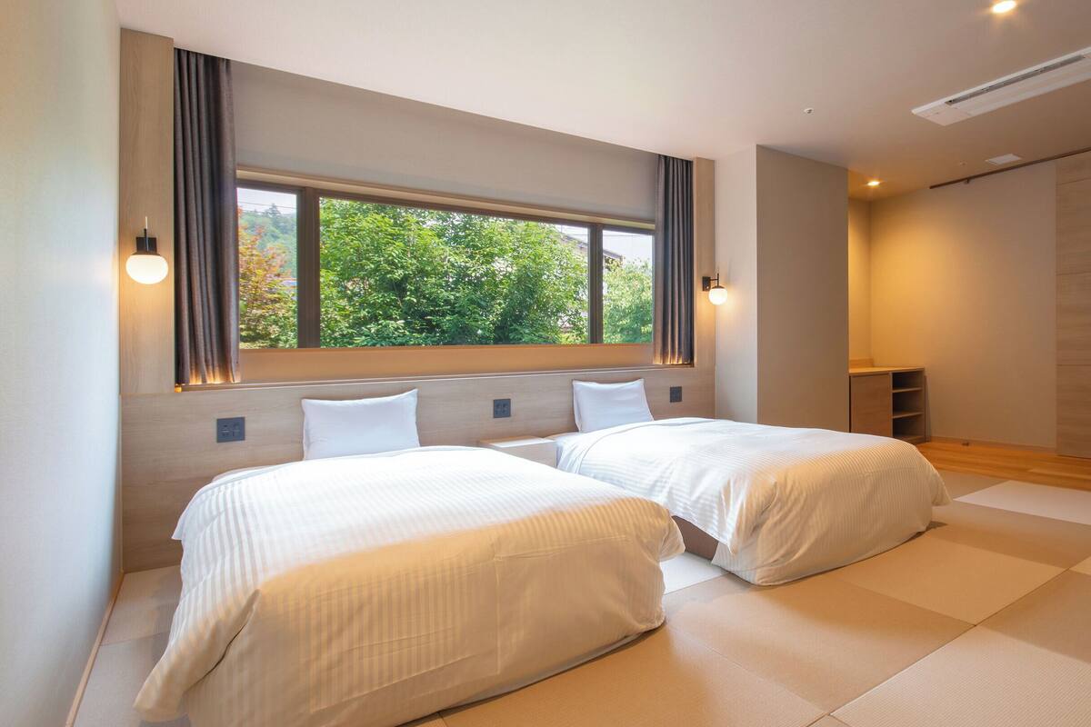 A modern hotel room with two beds, a large window with curtains, a built-in shelf, and a sliding door closet.