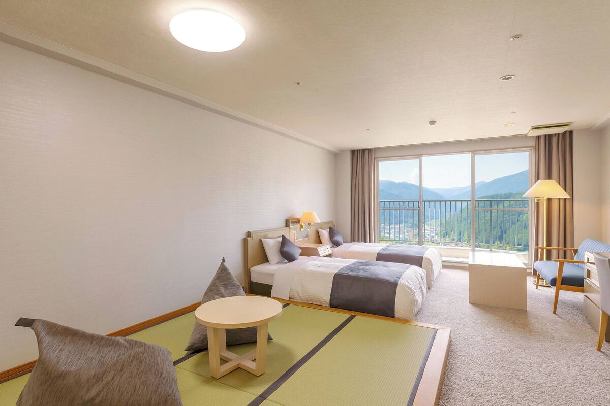A hotel room with two beds, a desk, a chair, a small table, and a view of mountains through large windows.
