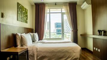 A hotel room with a large bed, a bedside table, a window with curtains, a wall-mounted air conditioner, and a view of the cityscape.