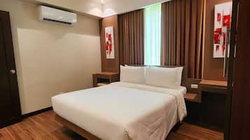 A hotel room with a large bed, wooden flooring, a wall-mounted air conditioner, and a window with curtains.