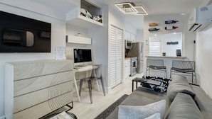 A modern living room with a sofa, a TV, a desk with a computer, a kitchen area, and a dining space with chairs.