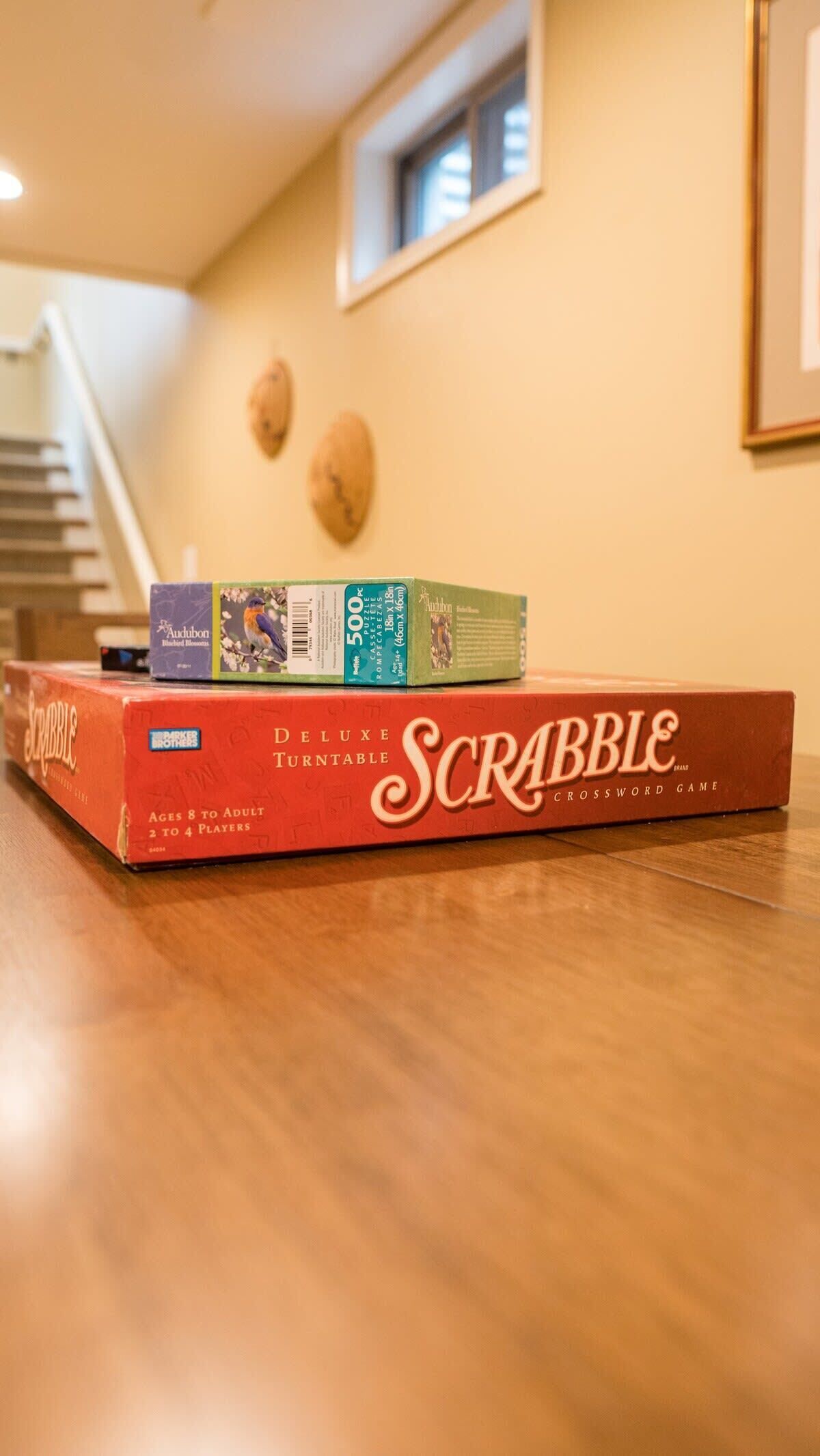 A Scrabble Deluxe Turntable game box on a wooden floor with a staircase and a small window in the background.