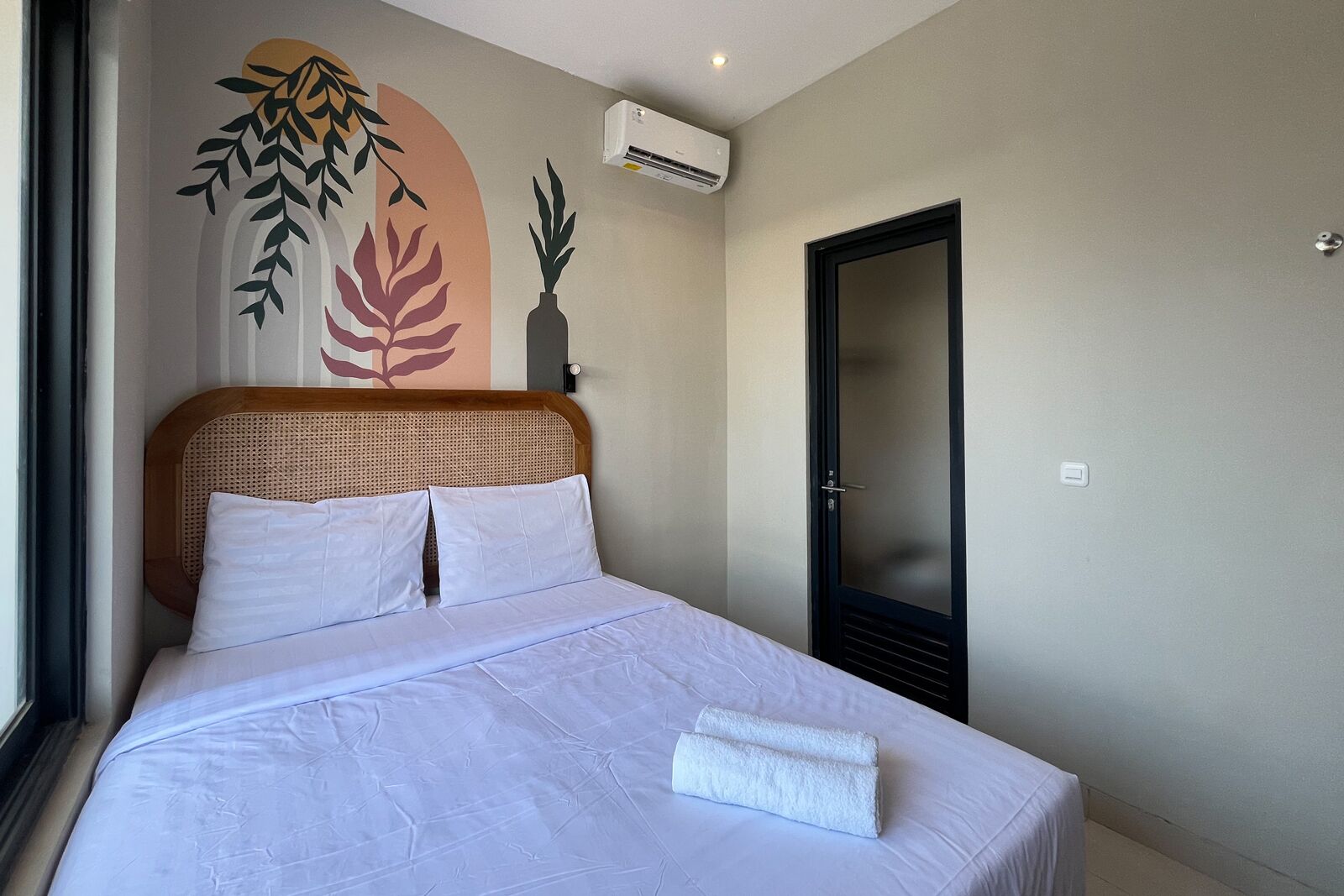 A neatly made bed with white linens, a rattan headboard, and a wall-mounted air conditioner.