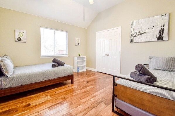 A room with two beds, wooden flooring, a white door, and a window with blinds.
