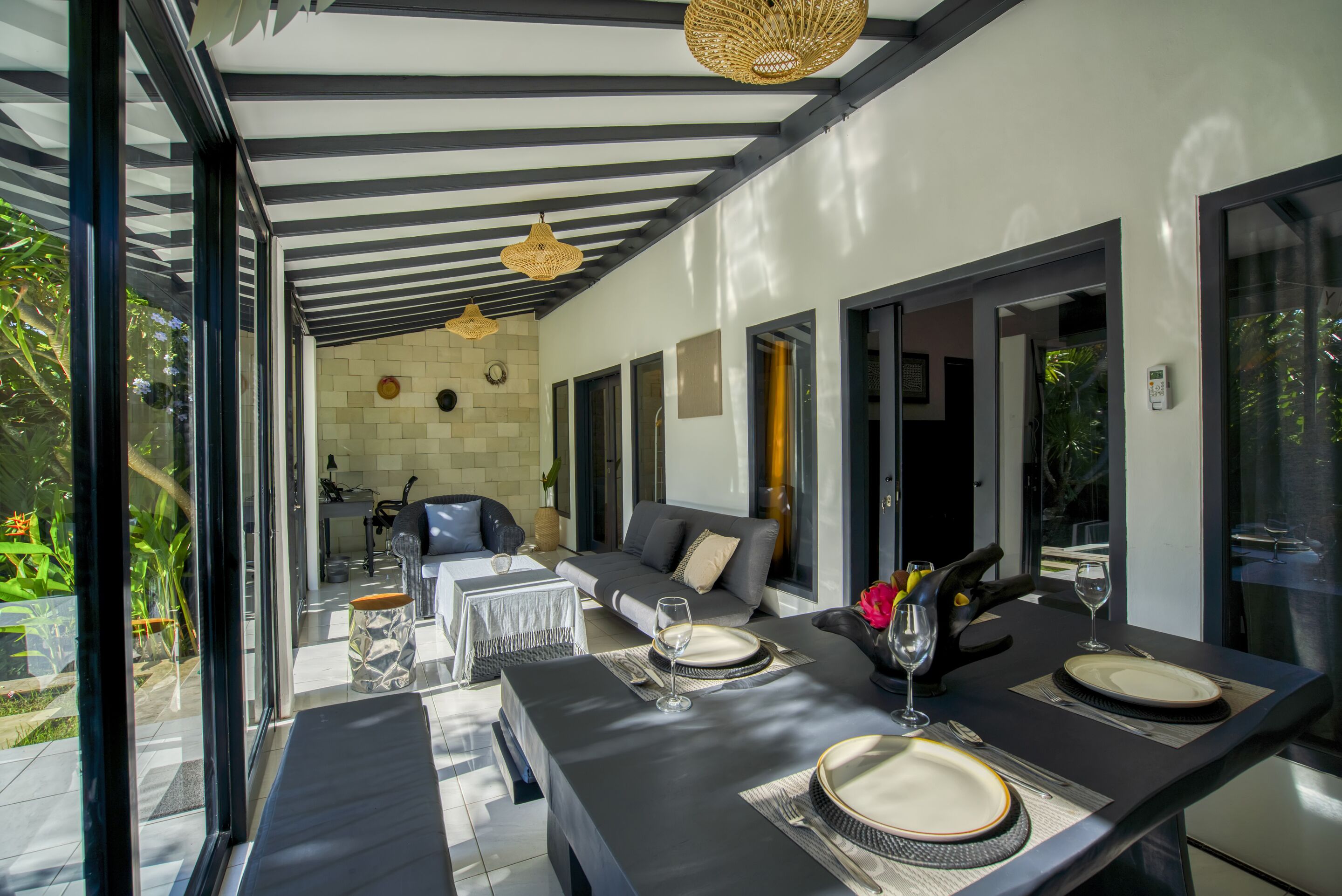 A modern dining area with a black table set for two, a sofa, and a view of the garden through large windows.
