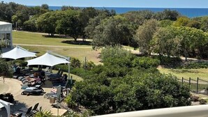 A view of a resort with a pool area, lounge chairs, and umbrellas. There are trees and a view of the ocean in the background. - The Pool Resort Kingscliff - One Bedroom Ocean and Pool View (4314) - Holiday Management Co. (Tweed)