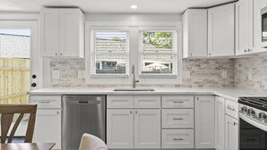 A modern kitchen with white cabinetry, a dishwasher, a stove, and a window with a view of the backyard.
