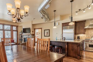Una cocina amplia con una isla grande, una mesa de comedor de madera y sillas. Hay una chimenea de piedra, un ventanal y una isla de cocina con taburetes de bar.