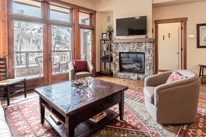 A living room with a fireplace, a television mounted above it, and a large window with a view of trees.