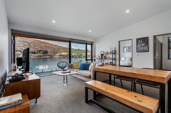 A modern living room with a large window overlooking a lake, a wooden dining table, and a cozy seating area.
