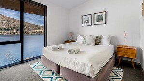 Quarto com uma janela grande, cama com travesseiros, mesa de cabeceira e abajur.
