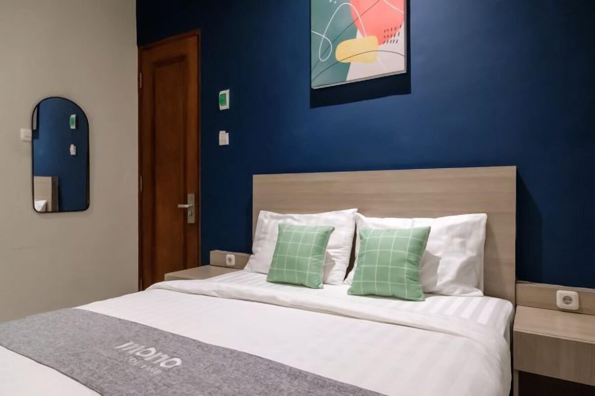 Une chambre d’hôtel avec un lit, une tête de lit en bois, un oreiller vert, un oreiller blanc, un plaid gris, un miroir, une porte et une œuvre d’art décorative accrochée au mur.