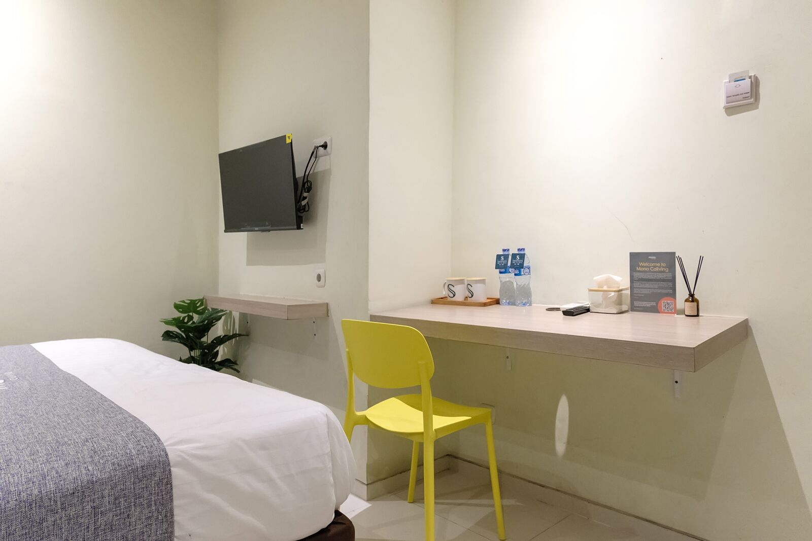 A hotel room with a bed, a desk, a chair, a TV mounted on the wall, a small plant, and a welcome sign.