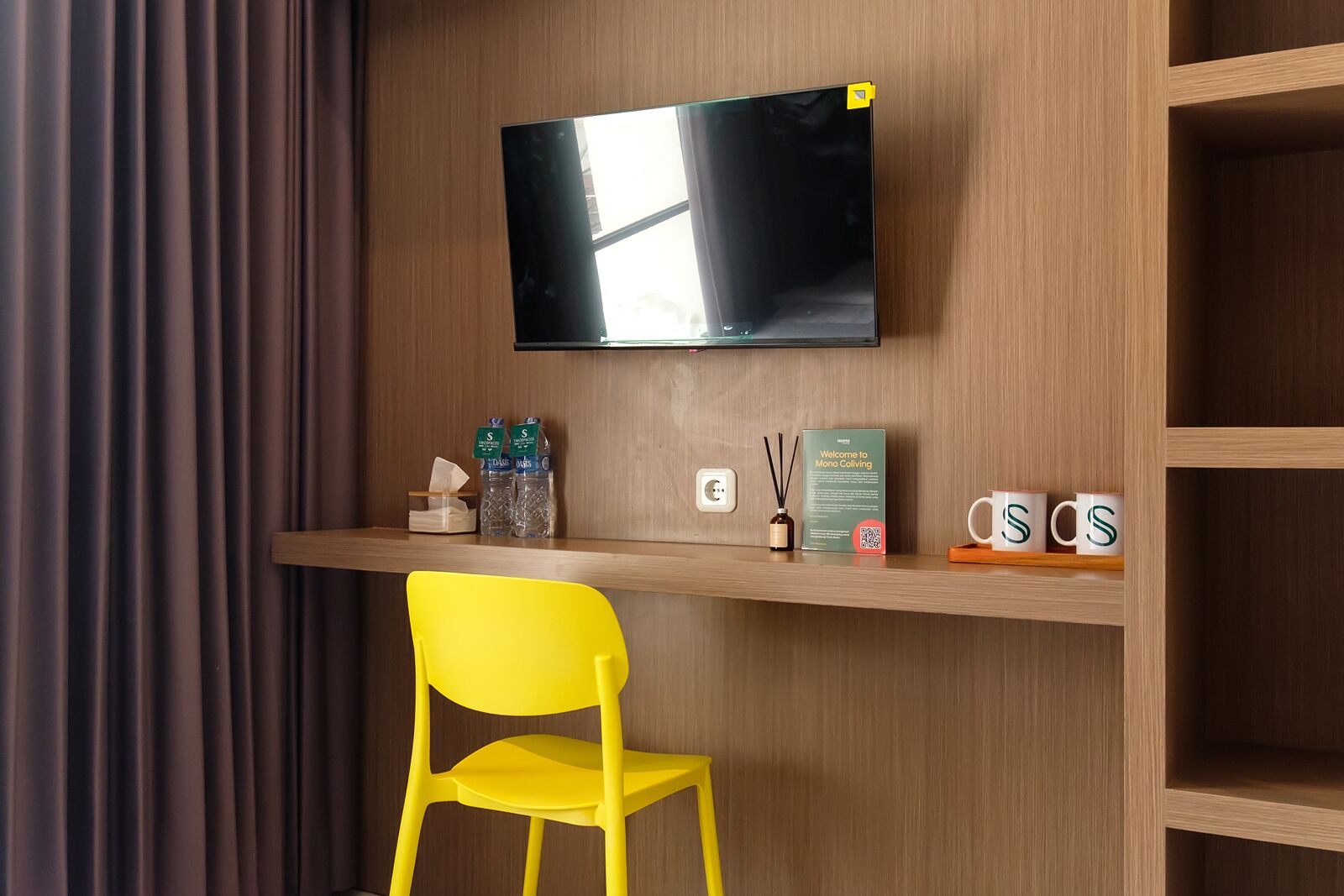 A modern hotel room with a wooden desk, a flat-screen TV mounted above it, a yellow chair, and a shelf with amenities including a tissue box, a bottle of water, a box of tissues, a diffuser, and two mugs.