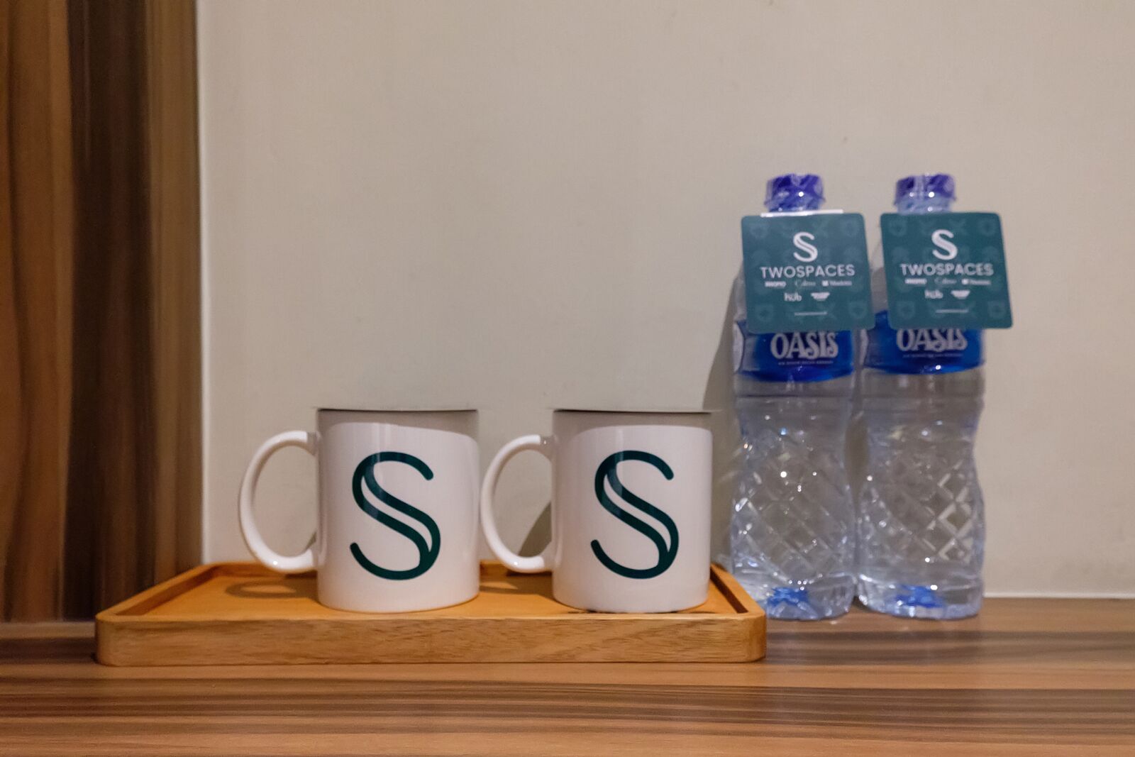 Deux tasses blanches ornées d’un logo « S » vert, posées sur un plateau en bois, à côté de deux bouteilles d’eau de la marque Oasis, portant l’étiquette « TWOPACES ».