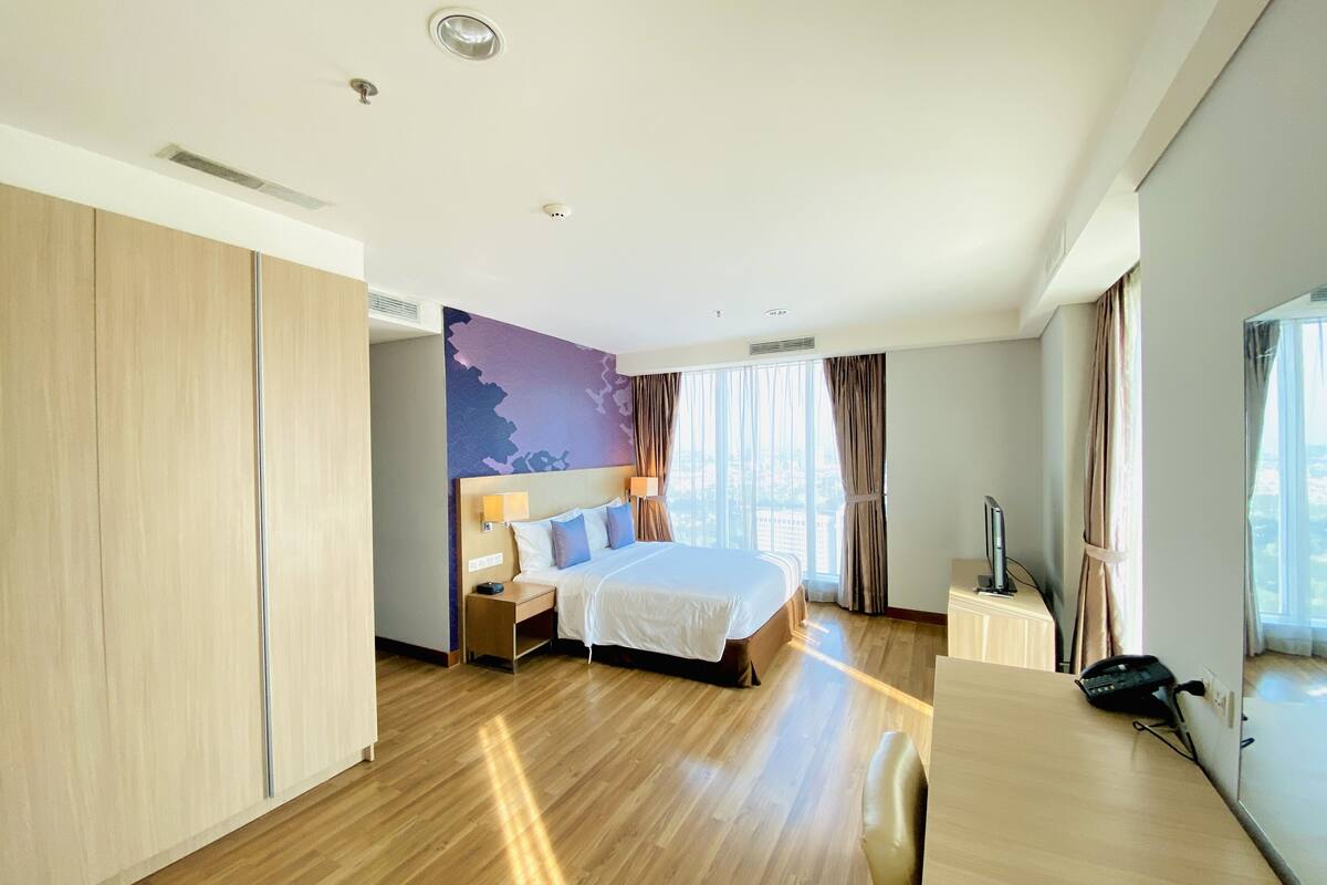 A modern hotel room with a large bed, wooden flooring, a desk with a telephone, and a view of the city through large windows.