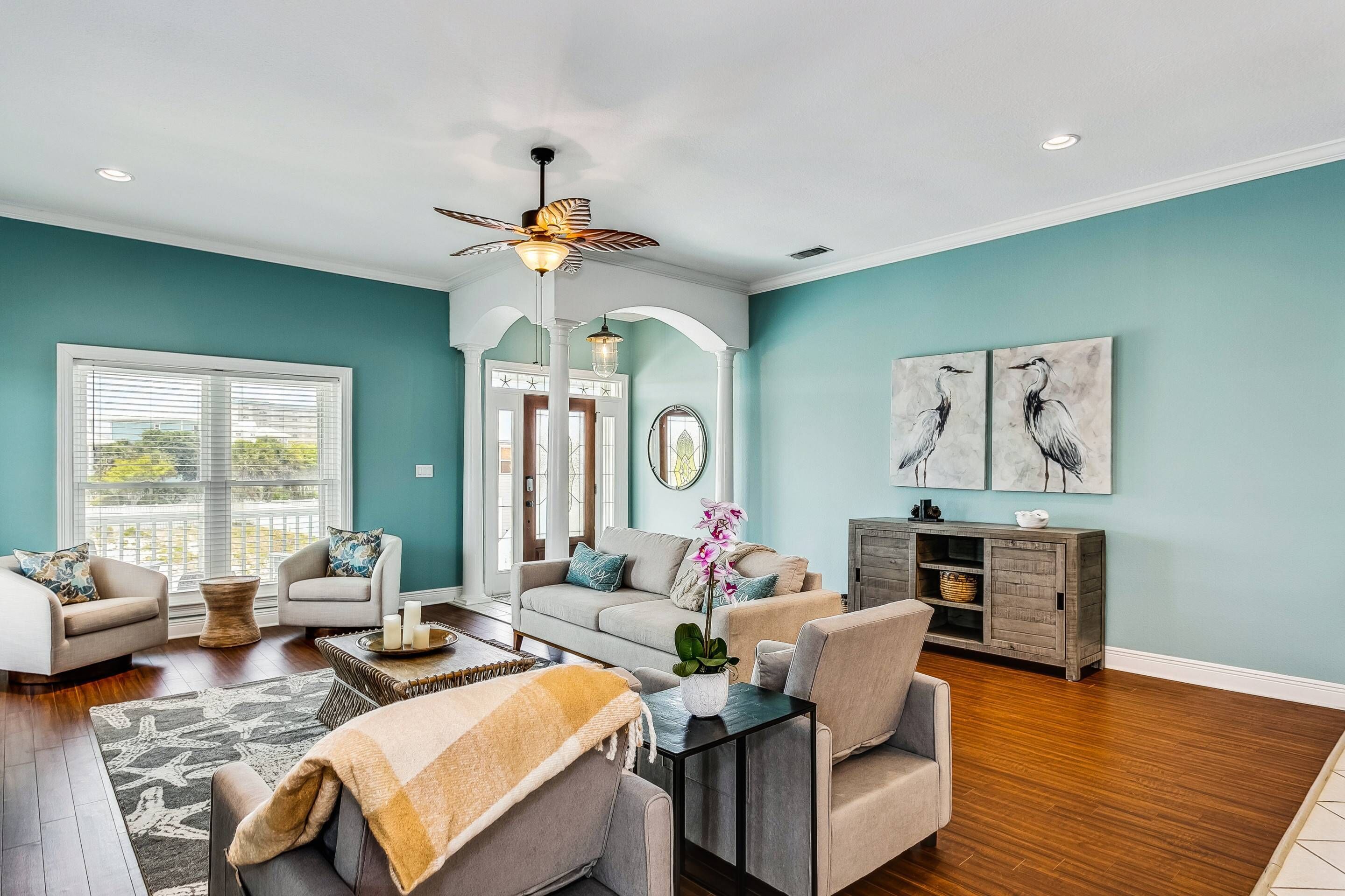 A living room with a teal wall, a ceiling fan, a large window, and a wooden floor.
