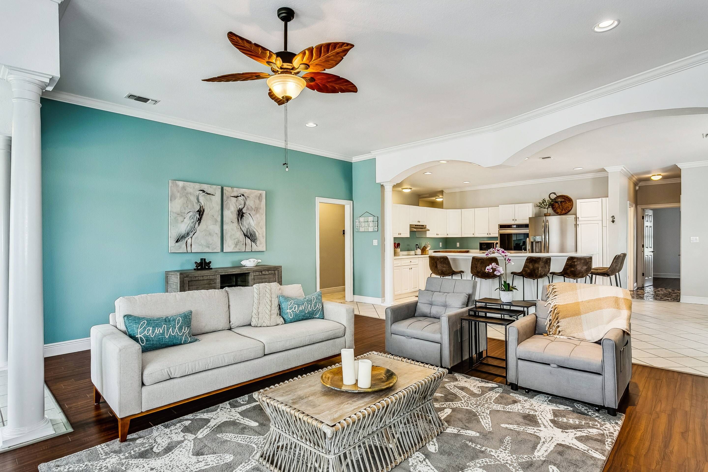 A spacious living room with a teal accent wall, a ceiling fan, and a modern kitchen in the background.