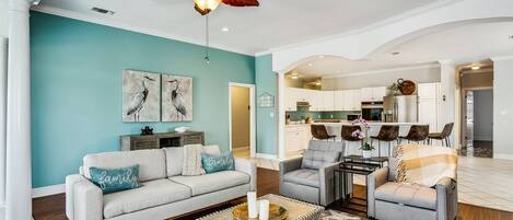A spacious living room with a teal accent wall, a ceiling fan, and a modern kitchen in the background.