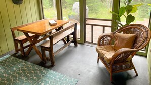 A wooden table with a bench, a wicker chair, and a view of a wooden deck and greenery through large windows.