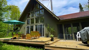 A wooden deck with outdoor furniture, a covered area with a green umbrella, and a large windowed building with a chimney.