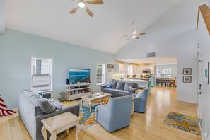 A spacious living room with a sofa, armchairs, a coffee table, and a TV. There is a kitchen in the background with a dining area.