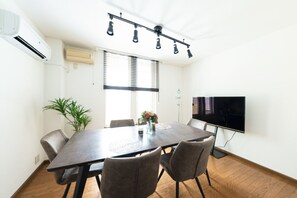 A modern dining area with a dark wooden table, four grey upholstered chairs, a potted plant, and a flat-screen TV mounted on the wall. - HONGO 11 HOTEL (Sapporo)
