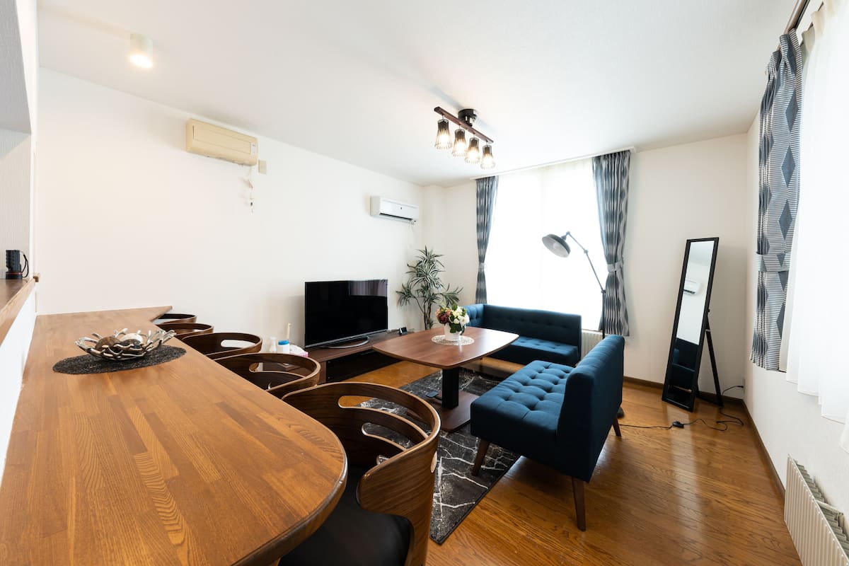 A modern living room with a wooden dining table, blue sofa, and a flat-screen TV.