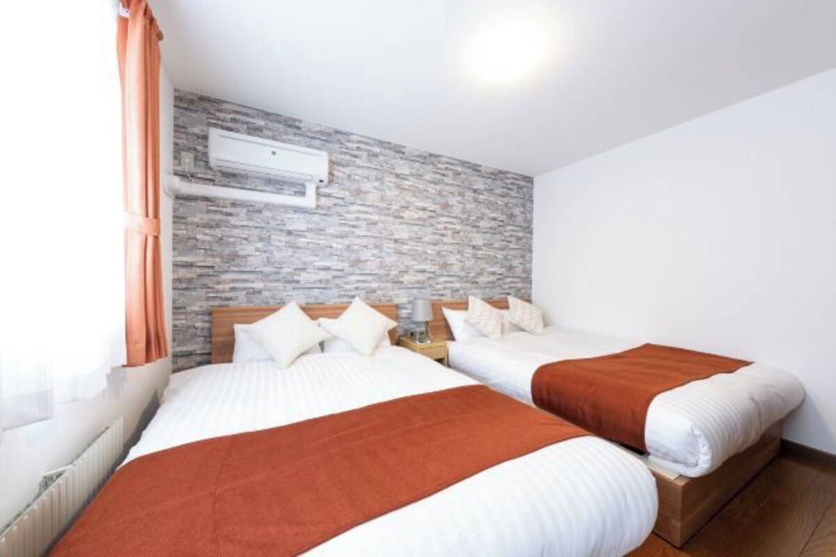 A hotel room with two beds, a stone-patterned wall, an air conditioning unit, and a window with curtains.